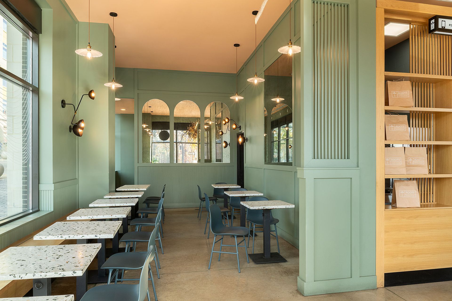 Roots Natural Kitchen interior with sage green walls, rows of terrazzo-topped tables, metal chairs, and light wood shelving.