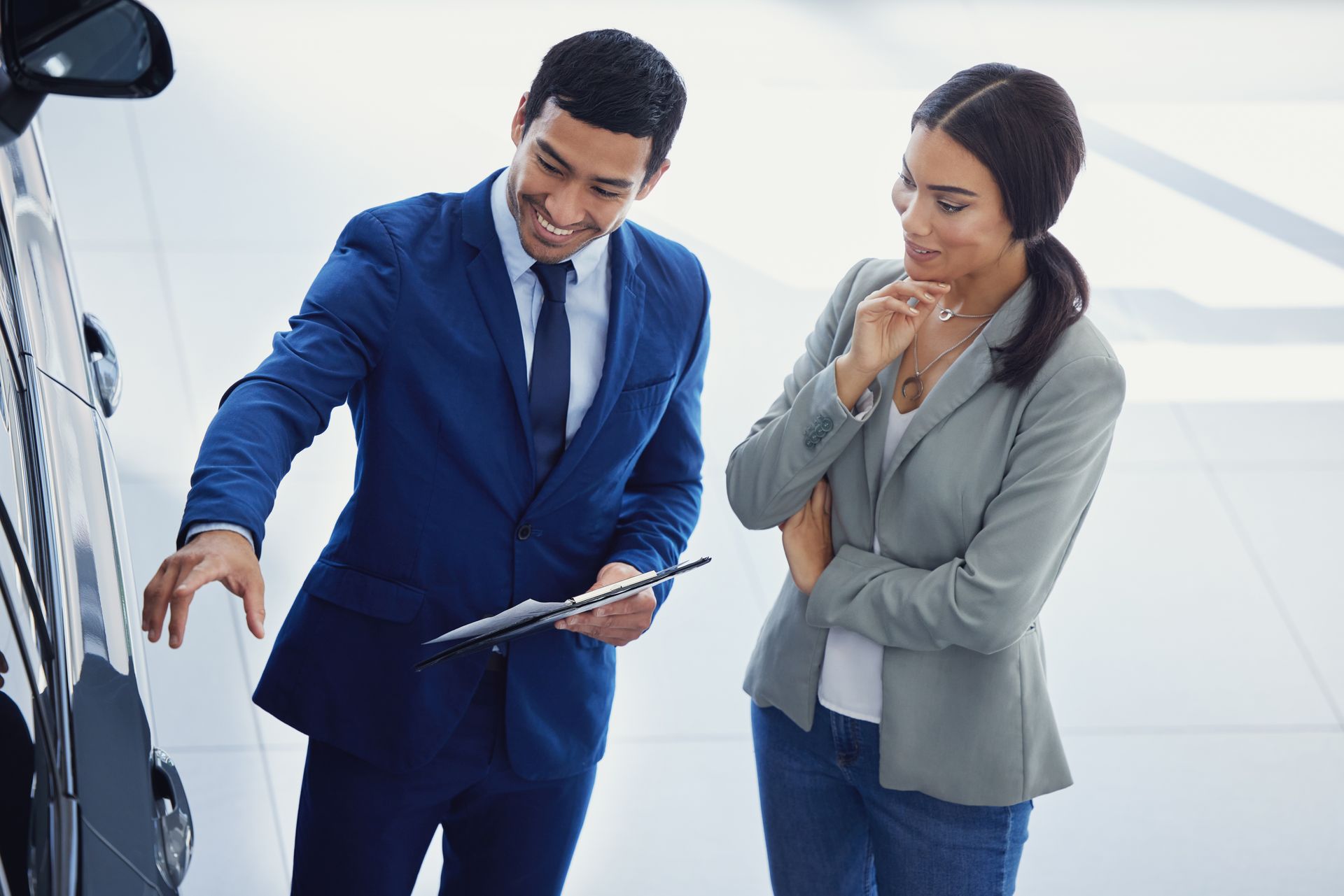 Car salesperson in a blue suit pointing at a black car, talking to a customer in a gray blazer and jeans inside a dealership.