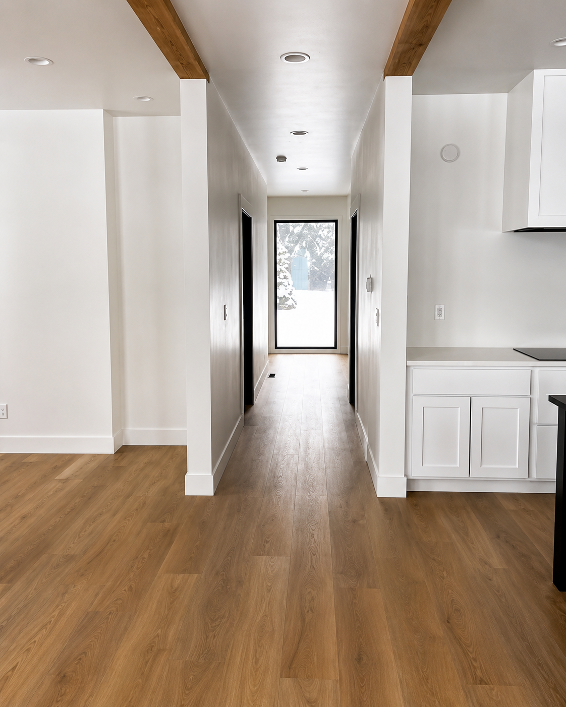 Empty hallway and kitchen with wood floors, white walls, exposed beams, and a glass door at the end.