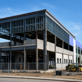 Steel-frame building under construction behind a chain-link fence on a clear day