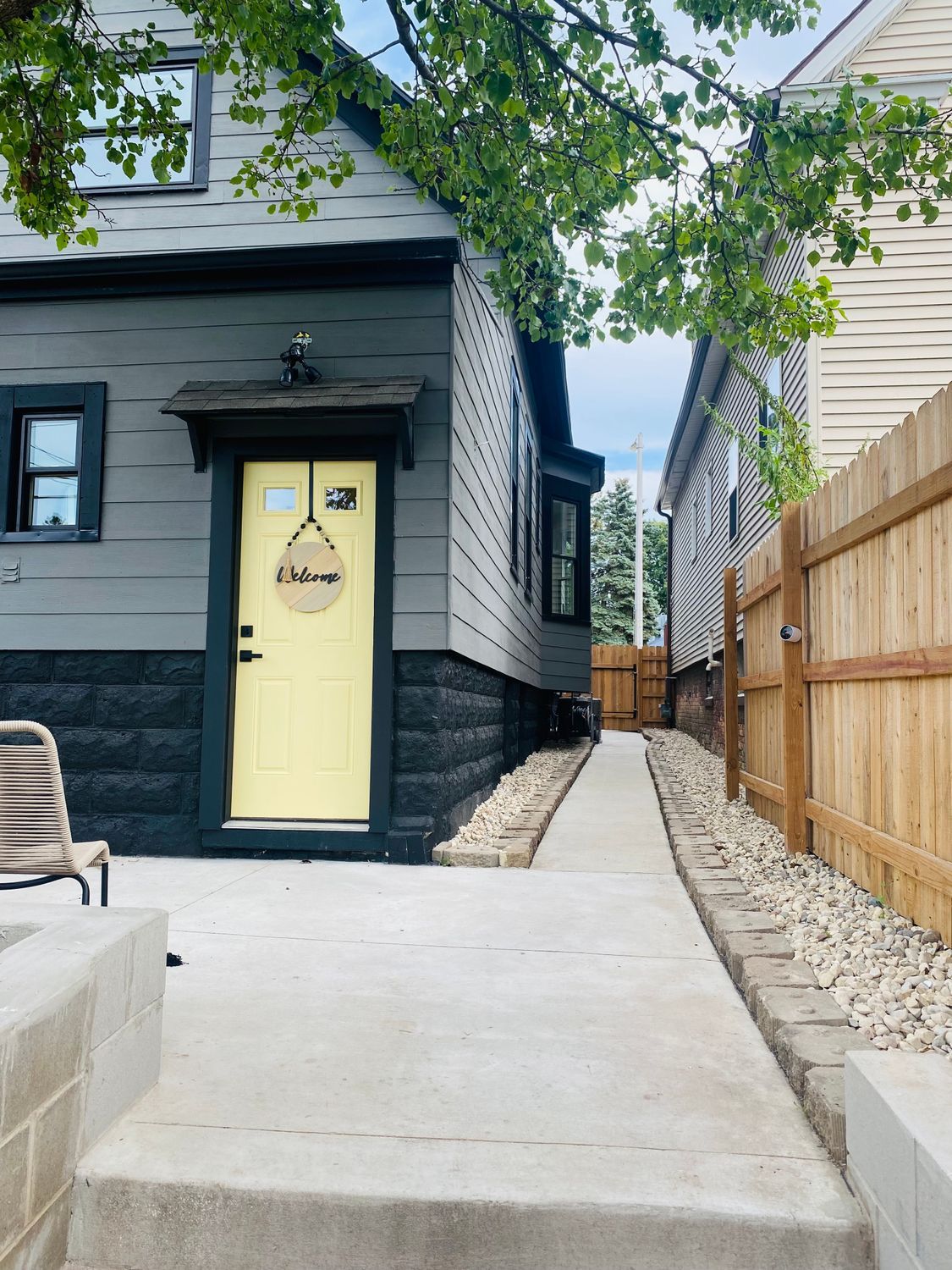 Narrow side walkway beside a gray house with a yellow door and wooden fence, leading to a backyard.