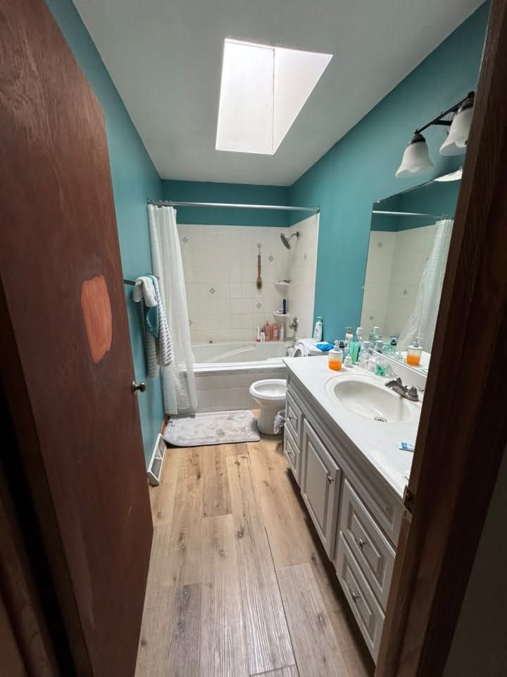 Narrow turquoise bathroom with skylight, double vanity, bathtub, and wooden floor viewed through an open door