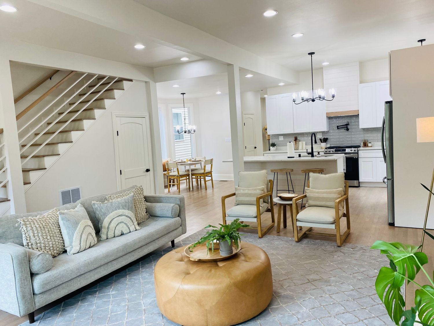 Bright open living room with gray sofa, tan ottoman, staircase, and adjoining kitchen/dining area