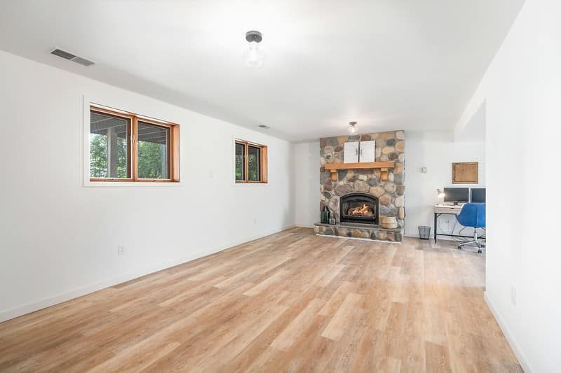 Empty living room with wood floors, white walls, two windows, and a stone fireplace.