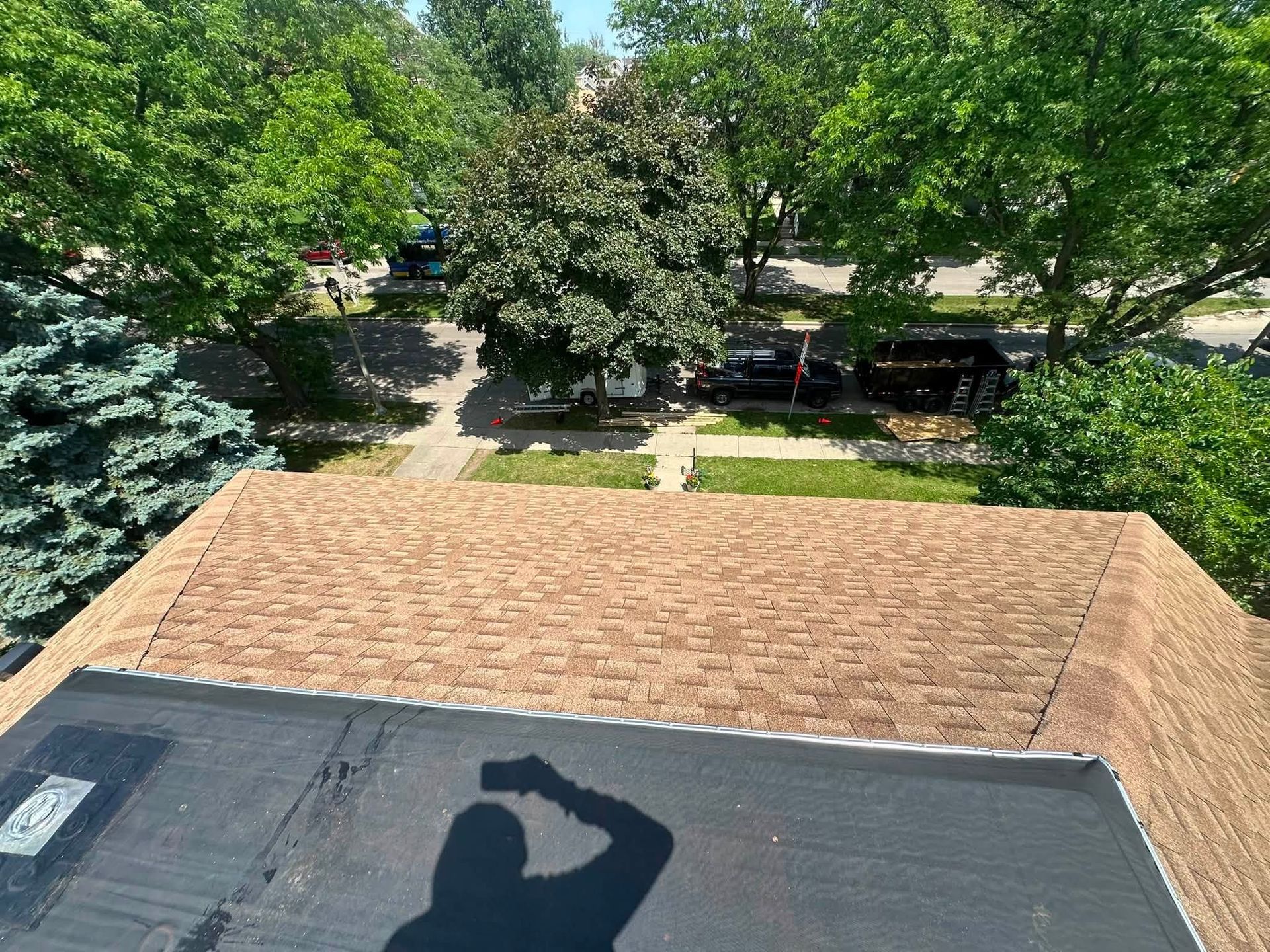View from a roof over shingles with a black tarp, overlooking trees and parked vehicles below