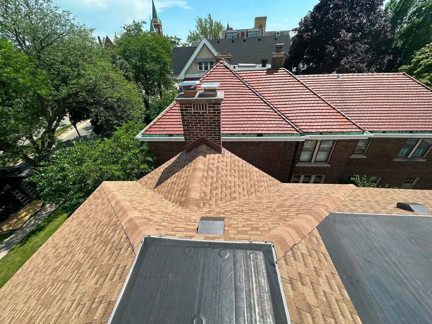 Rooftop view of a tan tiled roof with chimney, skylight, and neighboring houses under trees