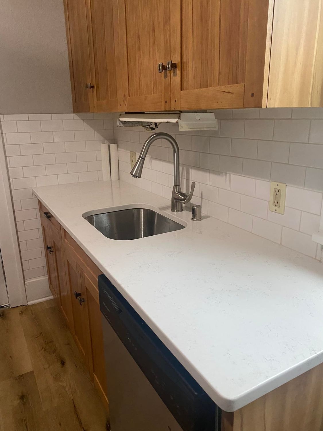 Small kitchen with white countertop, stainless sink, faucet, wood cabinets, and white tile backsplash.