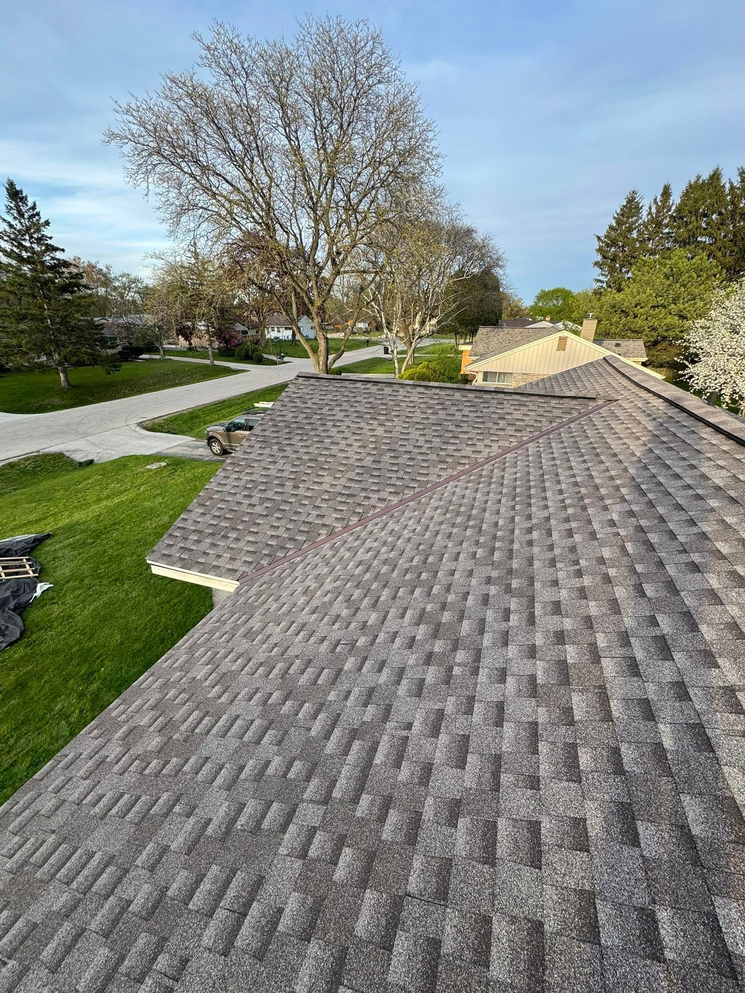 Gray shingle roof with trees and a driveway visible beyond it on a sunny day