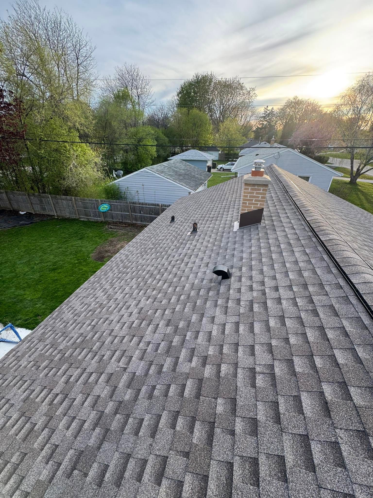 Shingled rooftop at sunset with vent pipes and trees in the background