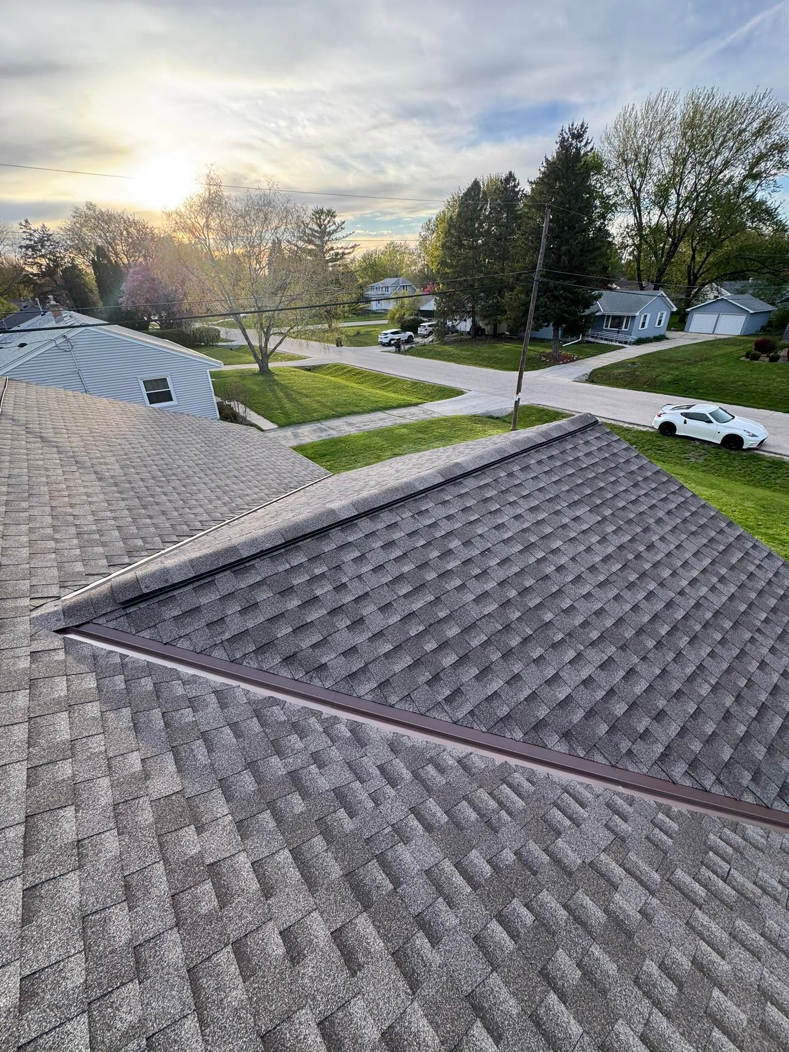 Rooftop shingles overlooking a suburban street at sunset, with trees and houses in the distance.