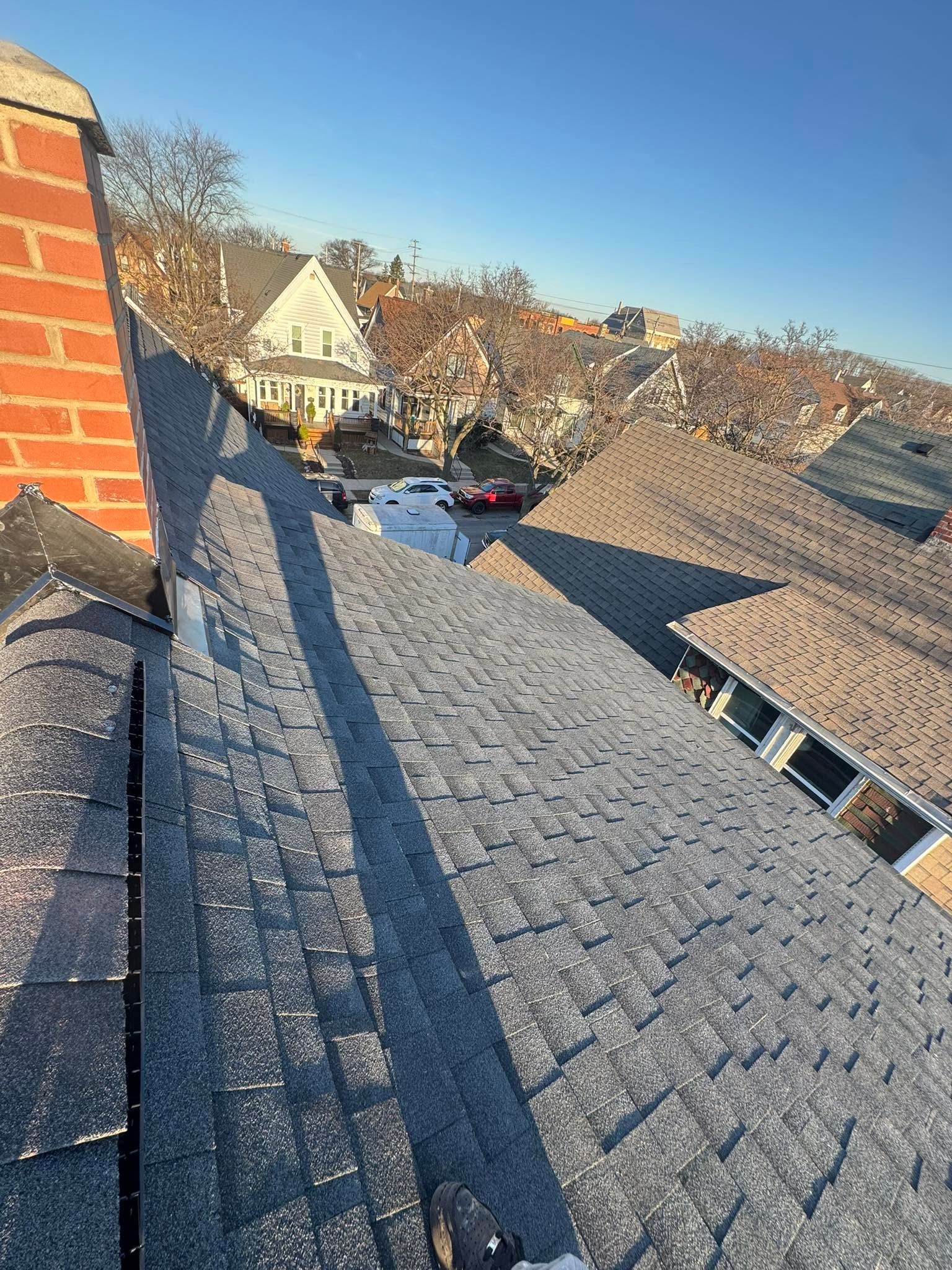 Rooftop view with gray shingles, brick chimney, and neighboring houses under a clear blue sky.