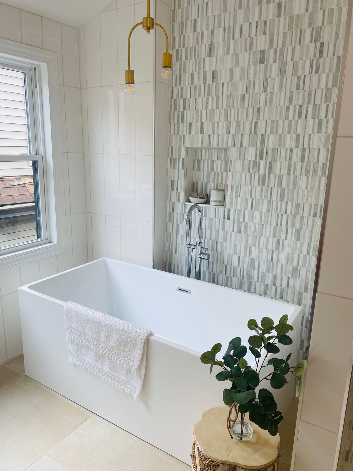 White freestanding bathtub beside a window, with a towel, gold shower, and mosaic tile wall.