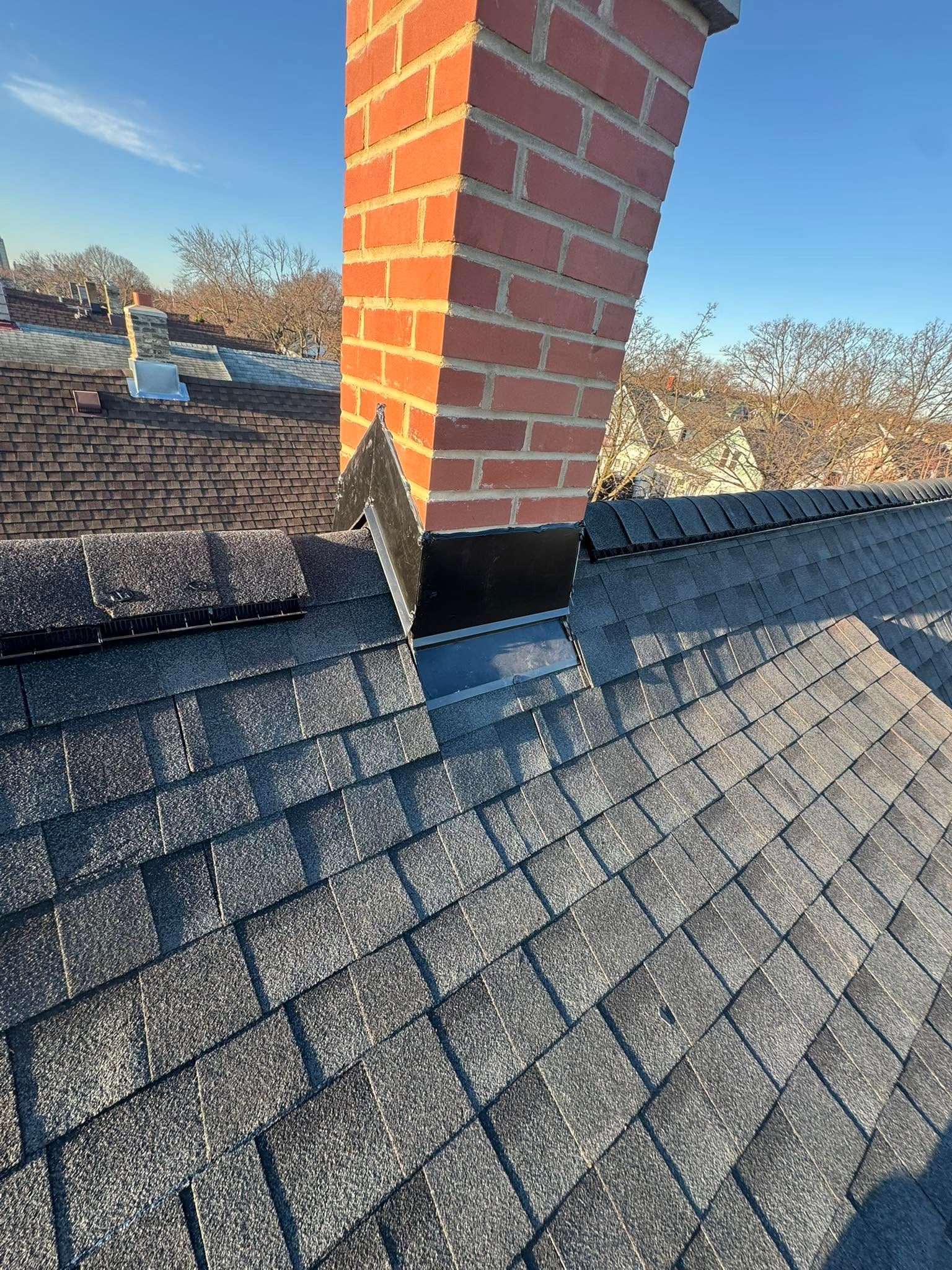 Brick chimney on a shingled roof with flashing, under a blue sky.