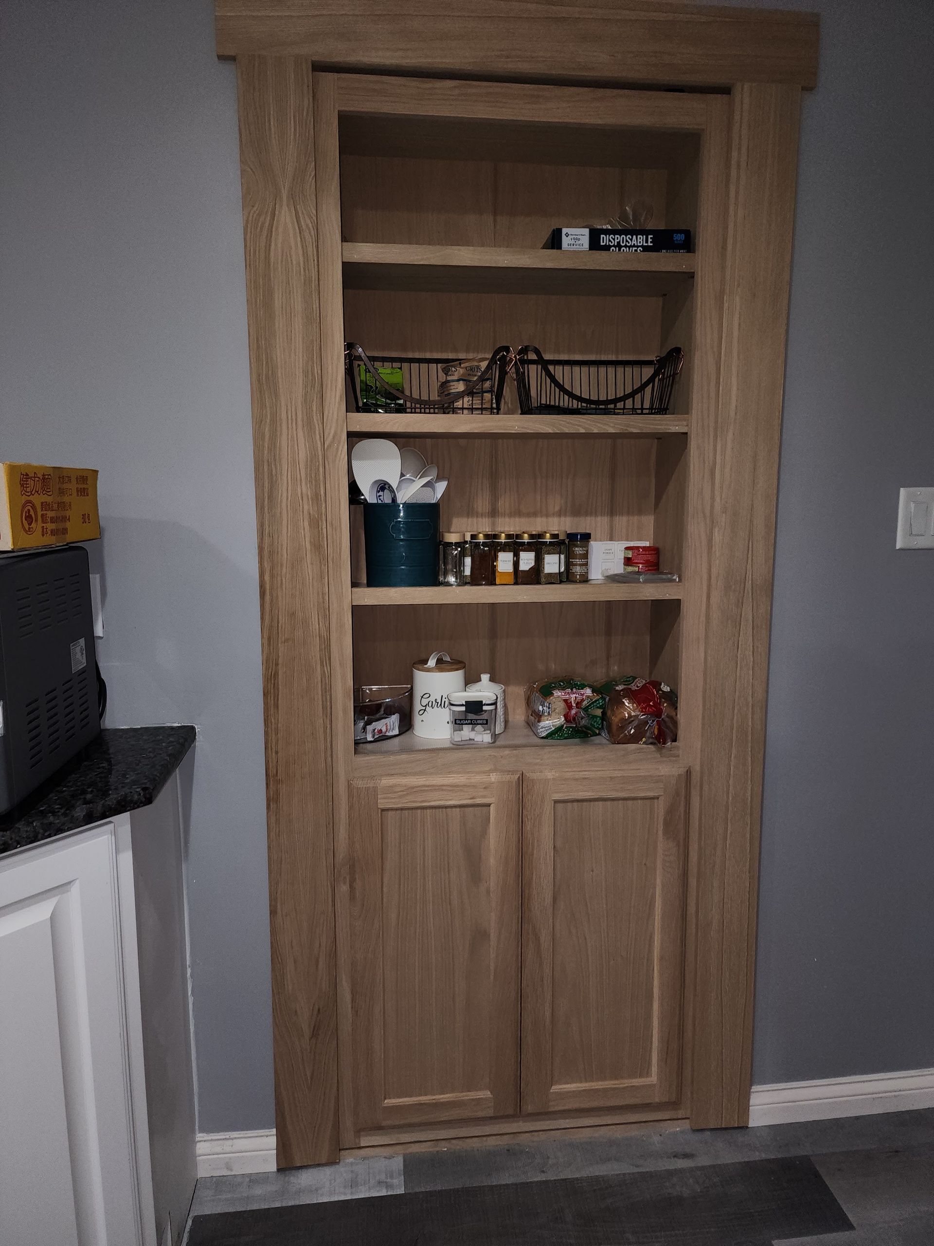 Wooden built-in pantry cabinet with shelves and glass doors, holding jars and kitchen items in a gray room
