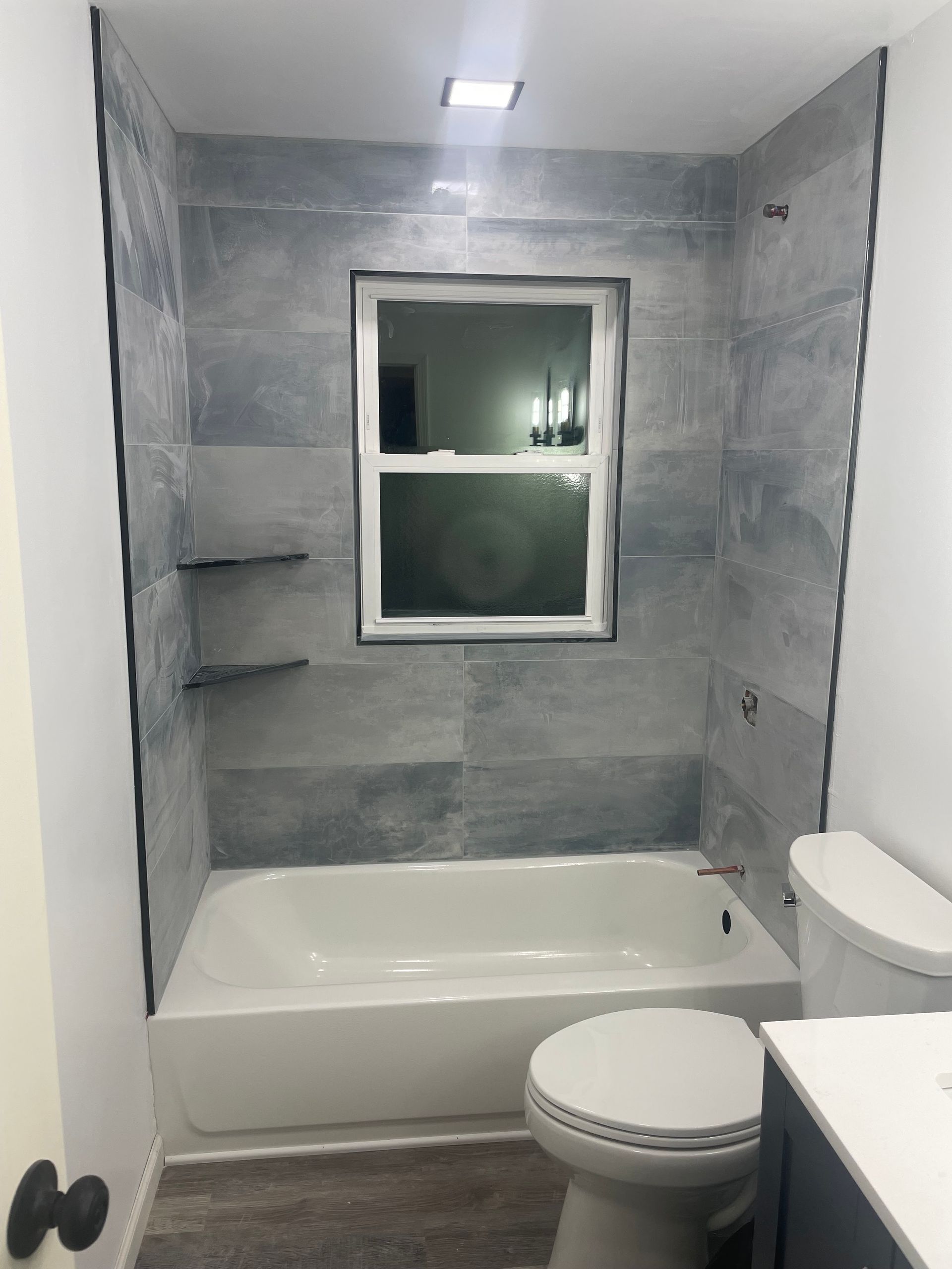 Small bathroom with gray tile, white tub, window, toilet, and vanity sink.