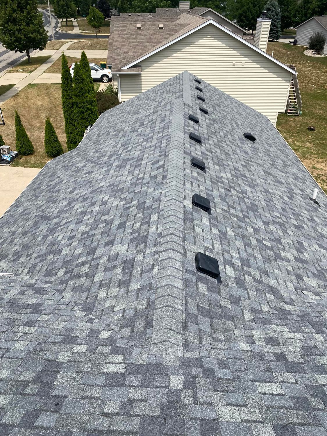 Gray shingled roof with roof vents, viewed from above toward a beige house and neighboring yards