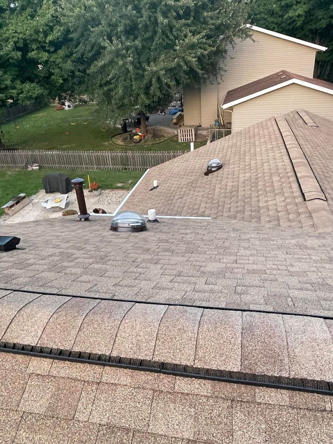 Rooftop with asphalt shingles and vents, seen over a suburban yard and house in the background