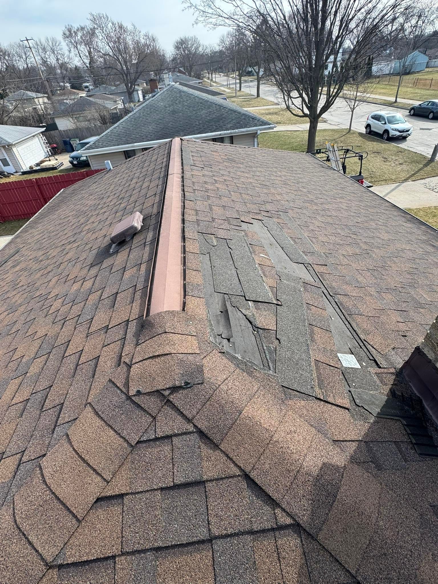 Damaged shingle roof with exposed ridge vent, viewed from above on a cloudy day