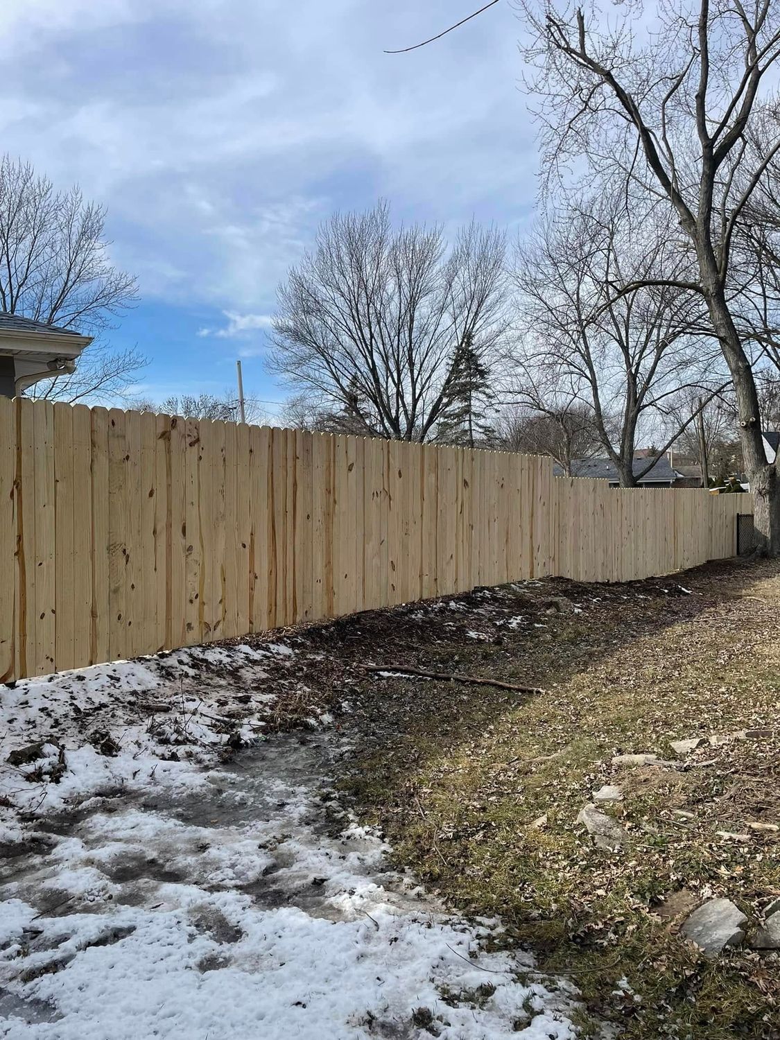 Wooden fence along a snowy backyard edge with leafless trees under a cloudy sky