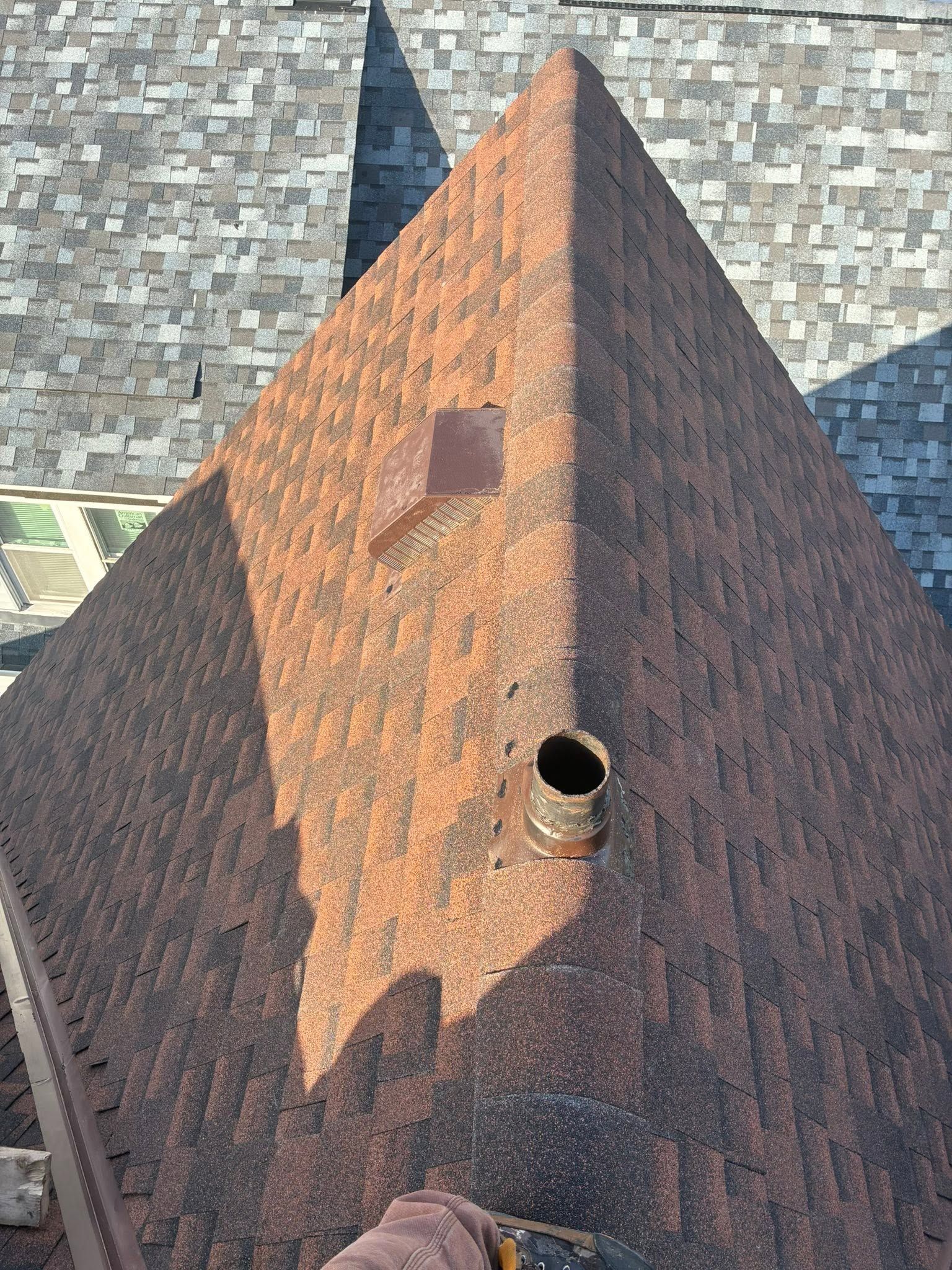 Steep shingled roof with a small pipe vent, lit by sunlight against a stone wall background