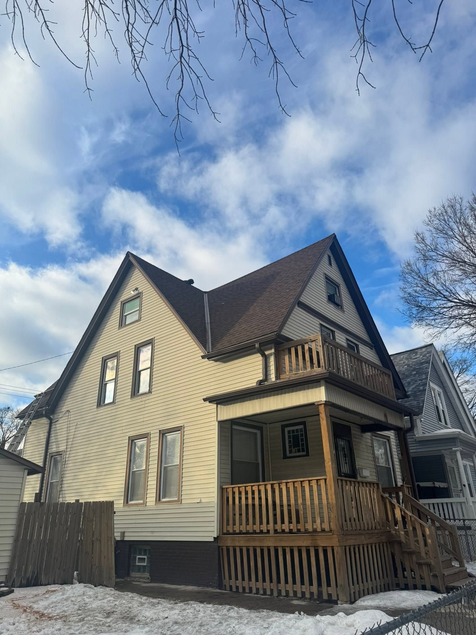 Victorian-style house with beige siding, dark trim, and a wooden porch under a partly cloudy sky.