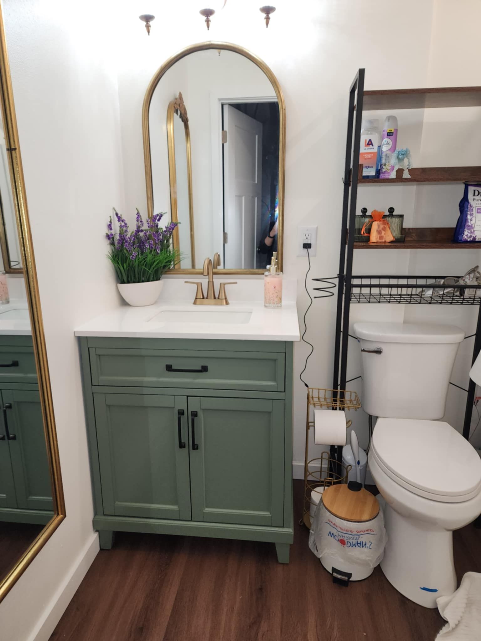 Bathroom vanity with arched mirror, green cabinet, white countertop, toilet, shelving, and woven basket on the floor