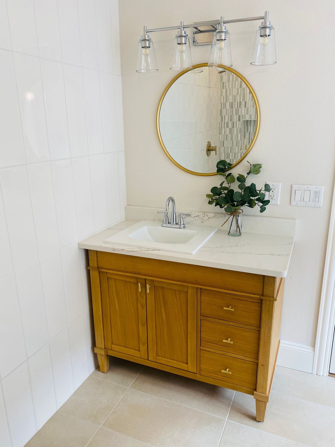 Bathroom vanity with round gold mirror, white sink, wooden cabinet, and vase of greenery
