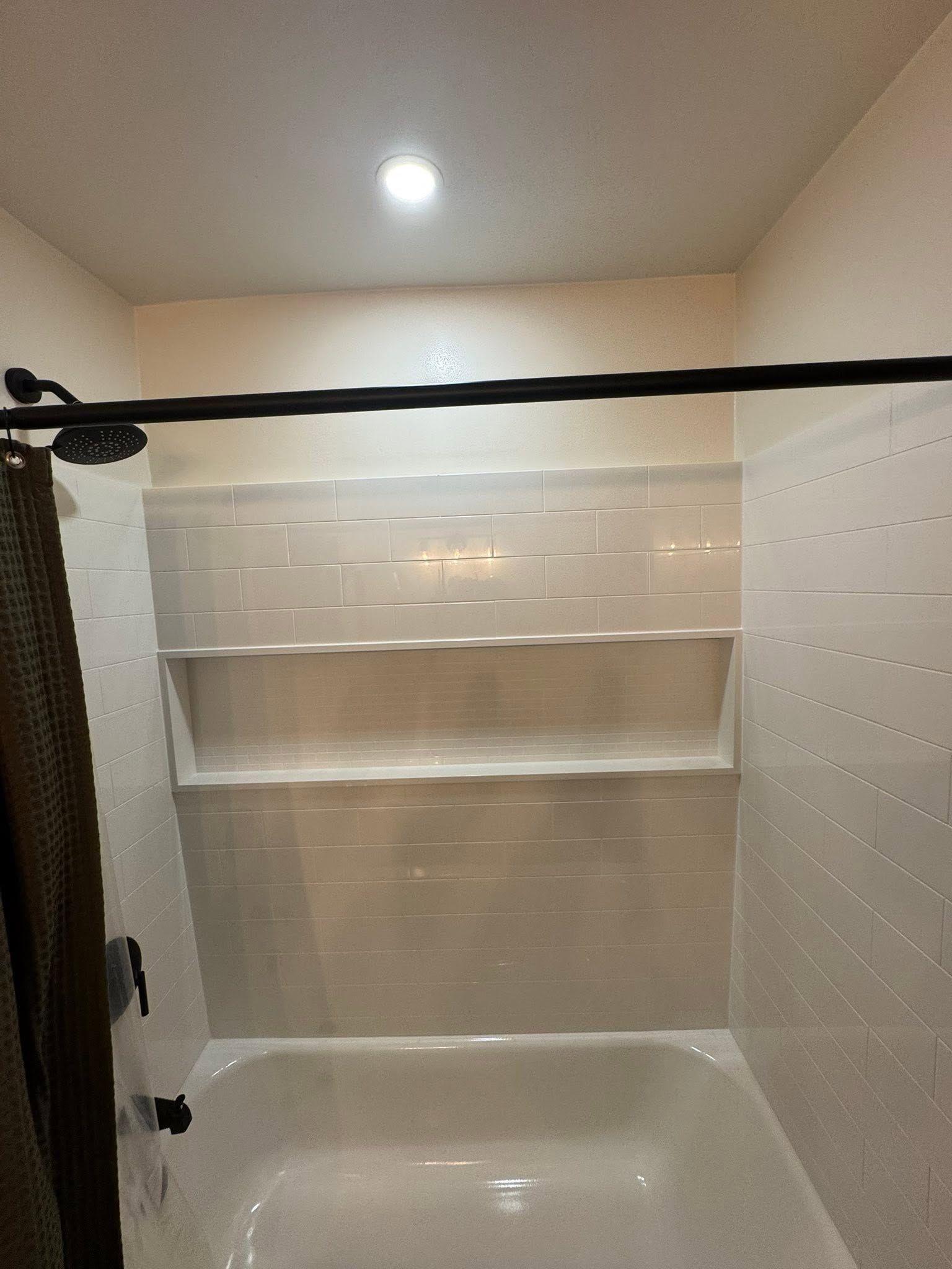 Small white tiled shower stall with a tub, built-in shelf, and overhead light