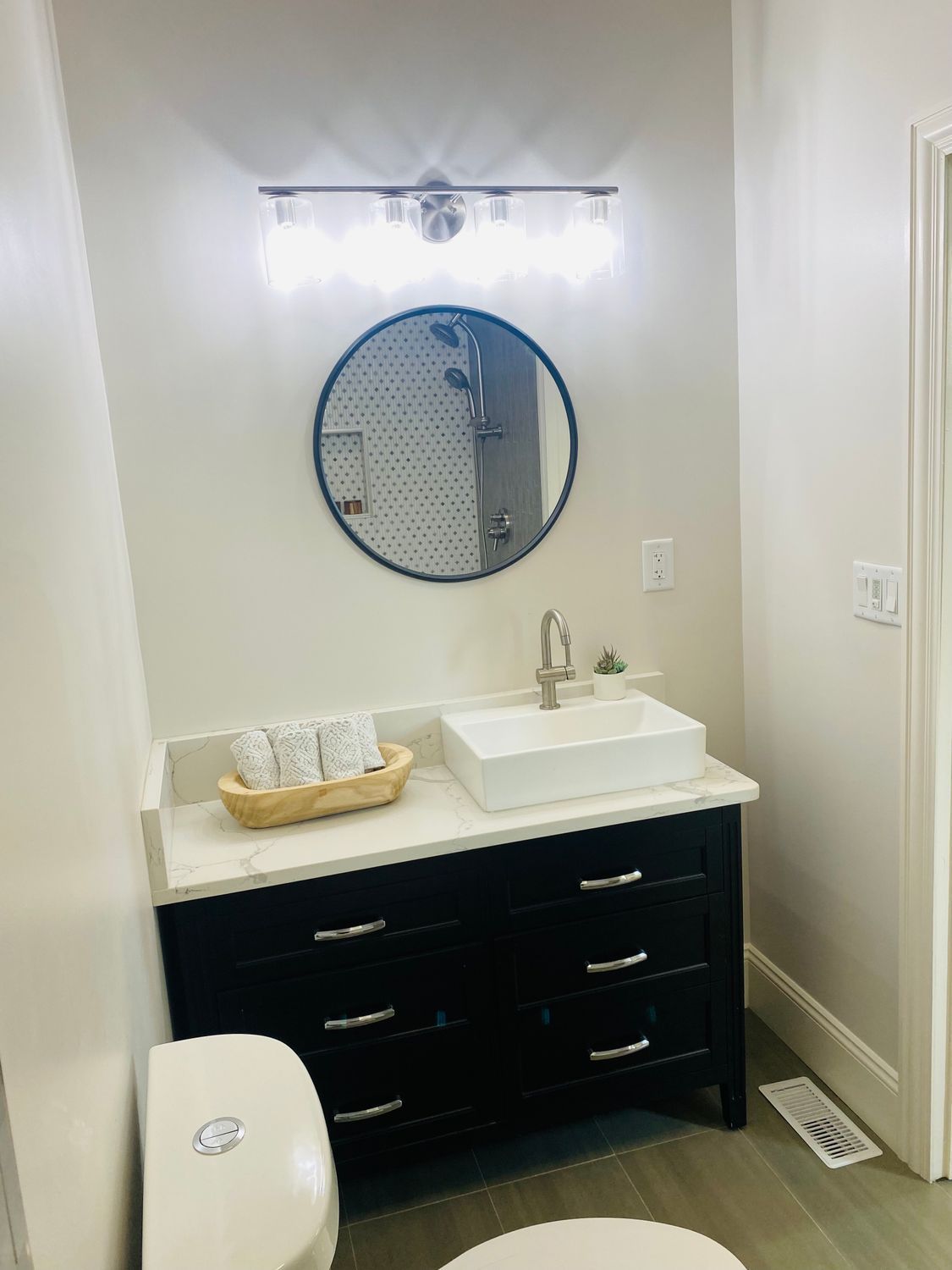 Small bathroom vanity with round mirror, white sink, dark cabinet, and wall light above.