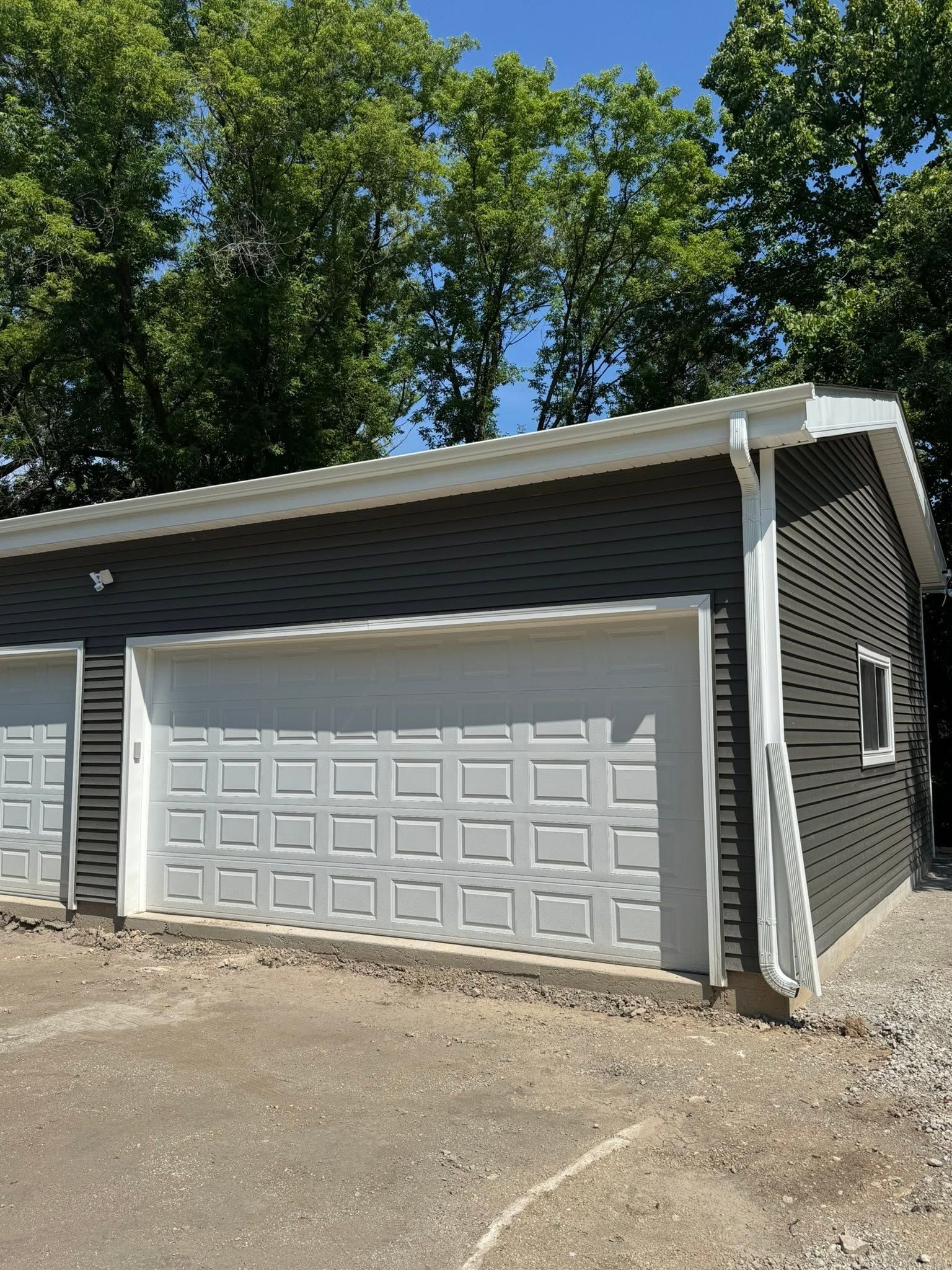 Detached gray garage with white door beside a driveway, trees in the background