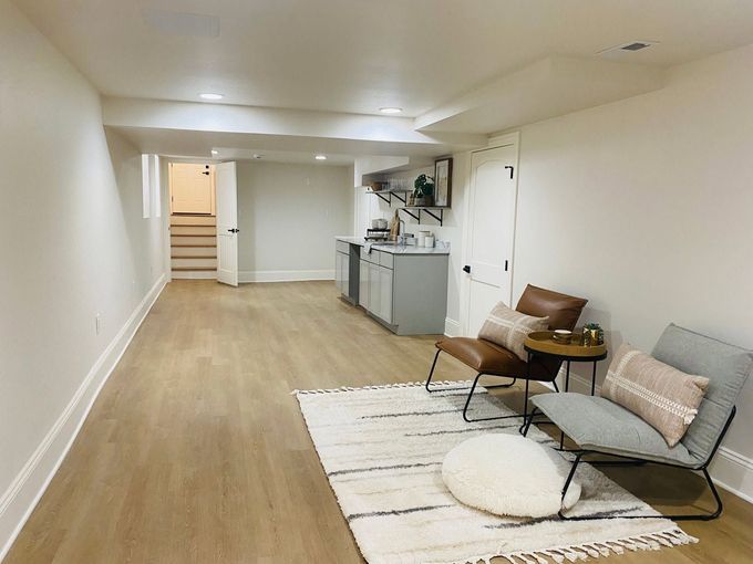 Spacious white basement living area with wood floor, small sofa, rug, and kitchenette at the far end.