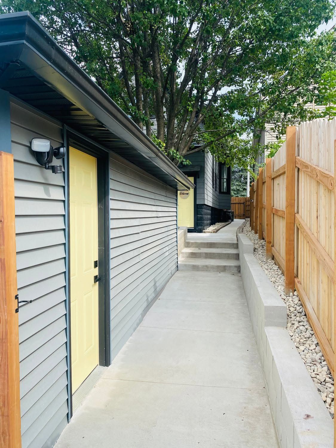 Narrow concrete walkway beside a gray garage and wooden fence, leading uphill past trees.
