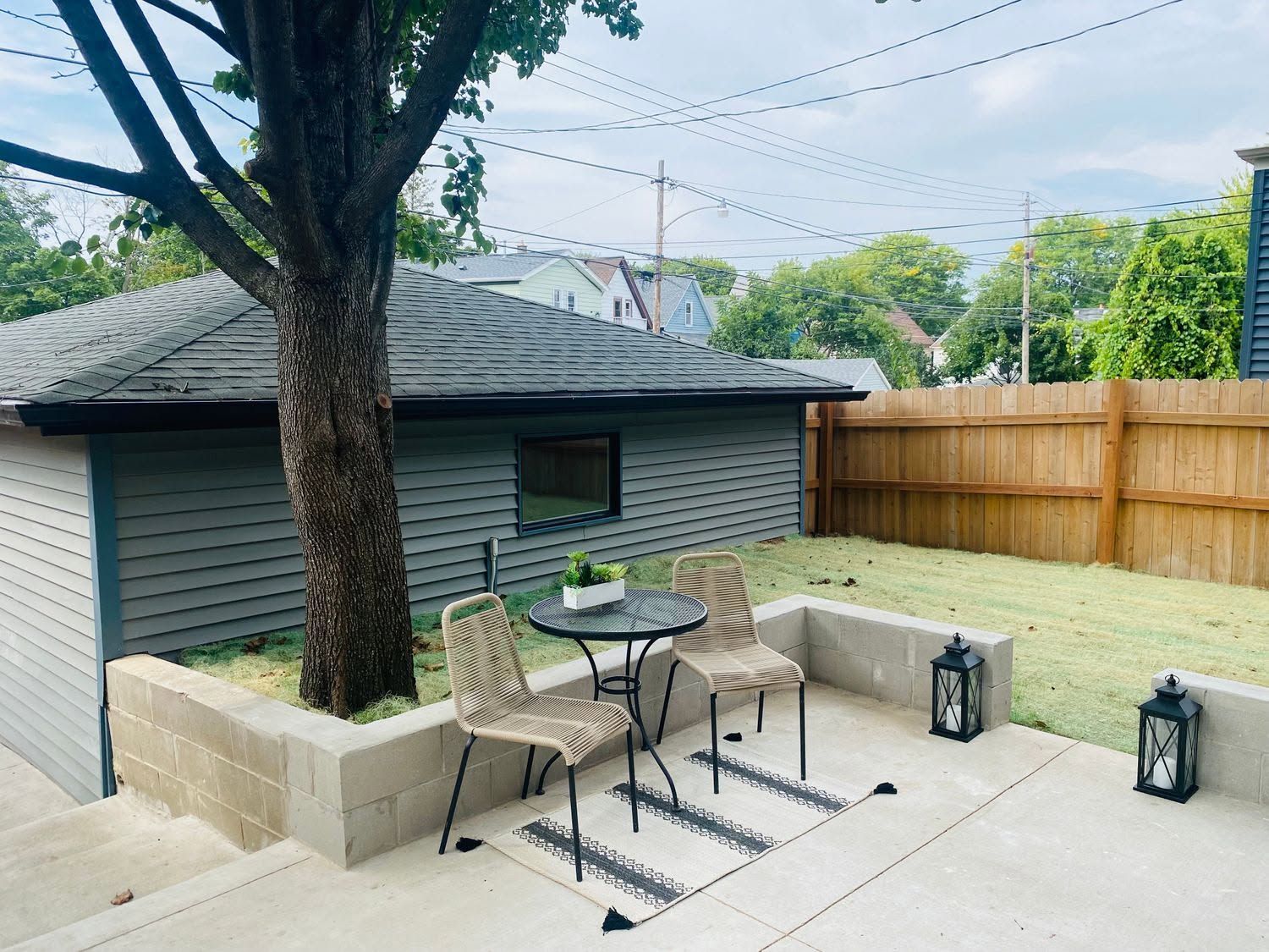 Backyard patio with table and chairs beside a small shed, tree, and wooden fence