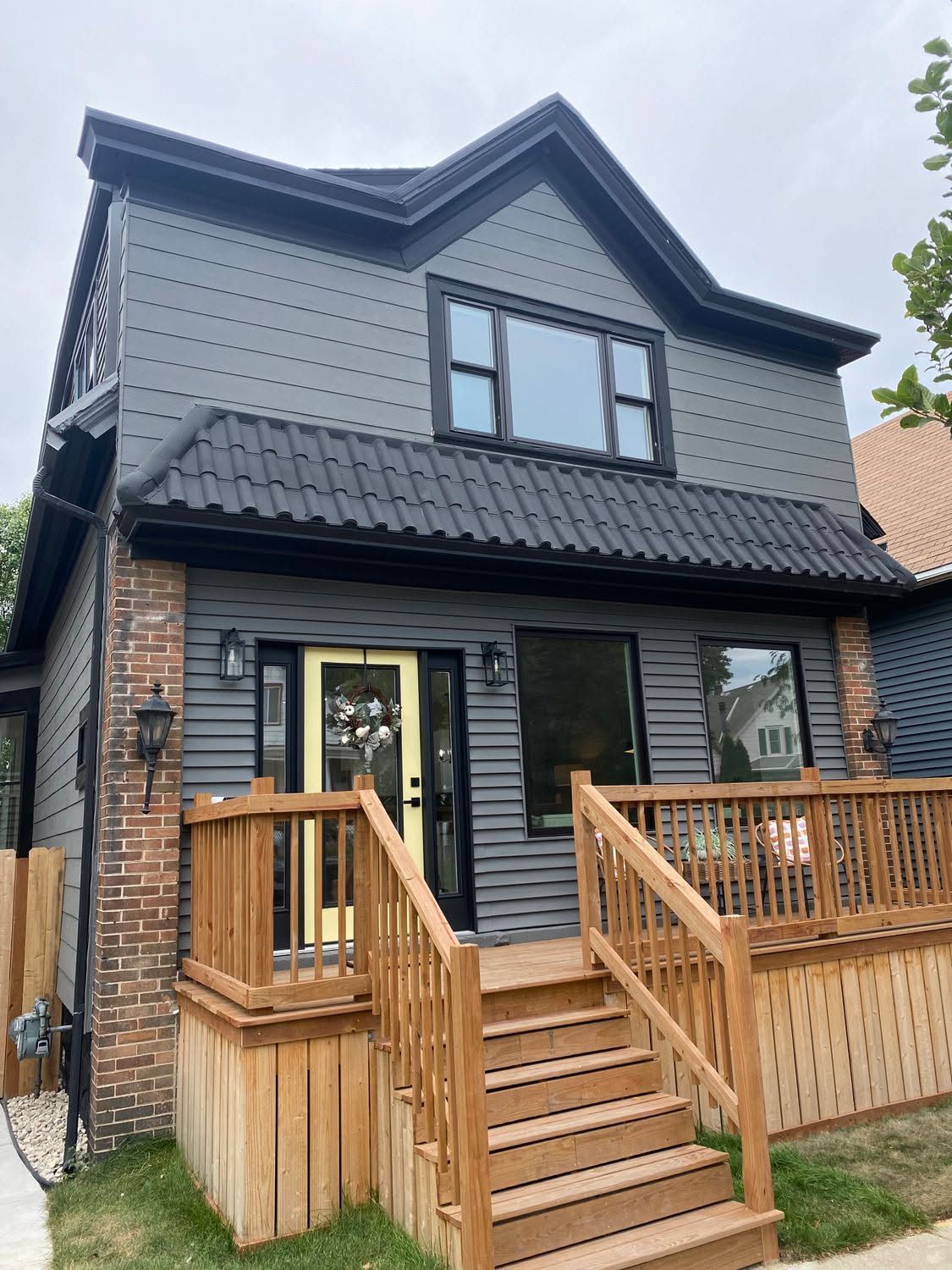 Two-story gray house with brick accents, black roof, and wooden front porch with stairs and railing.