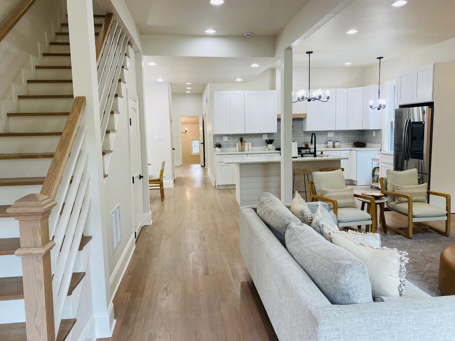 Bright open-concept living room and kitchen with hardwood floors, white cabinets, staircase, and gray sofa