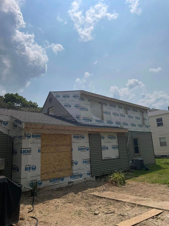 Partially built house with white wrap, boarded windows, and a dirt yard under a bright cloudy sky