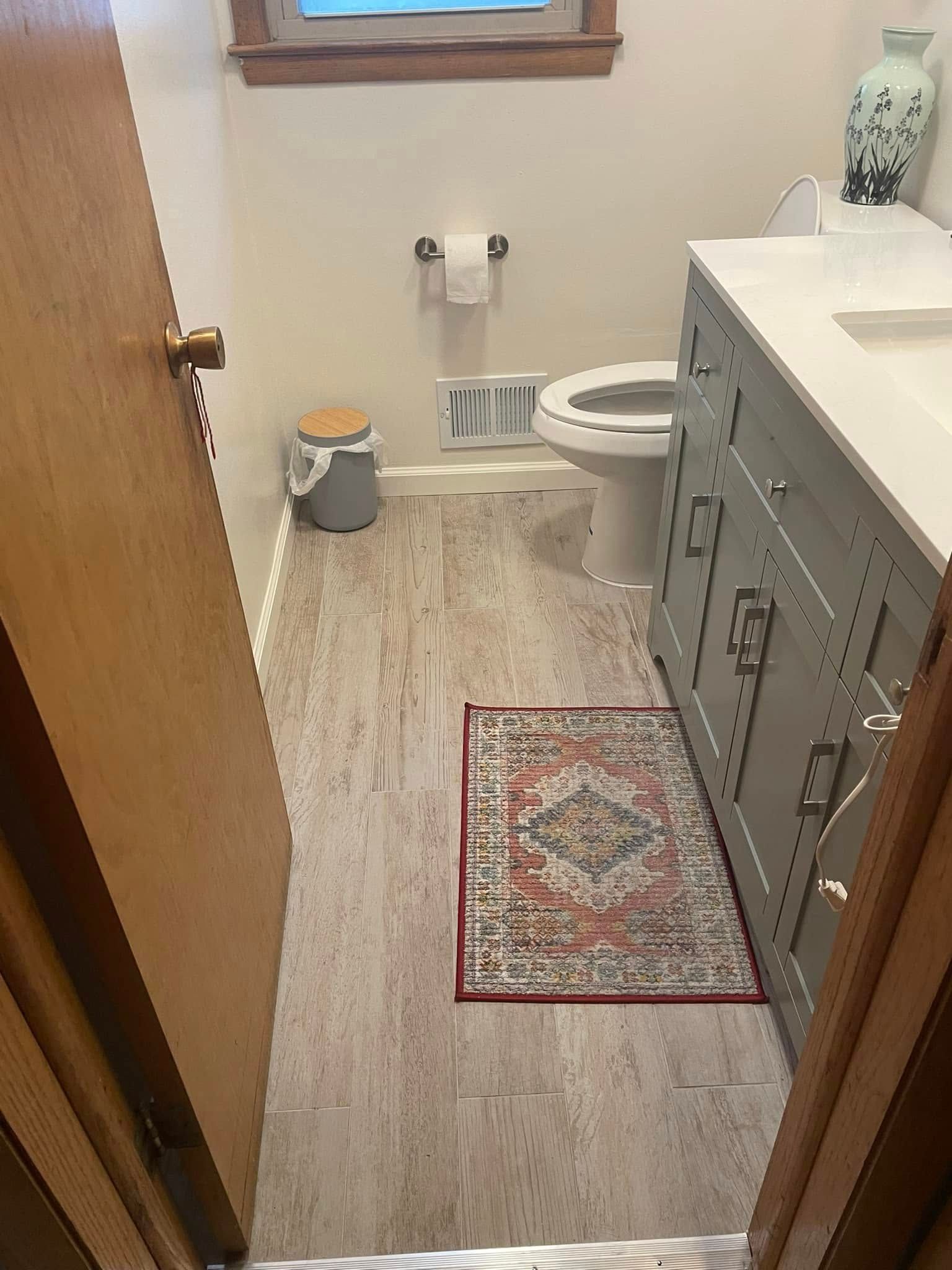 Small bathroom with open door, toilet, window, trash can, gray vanity, and red patterned rug.