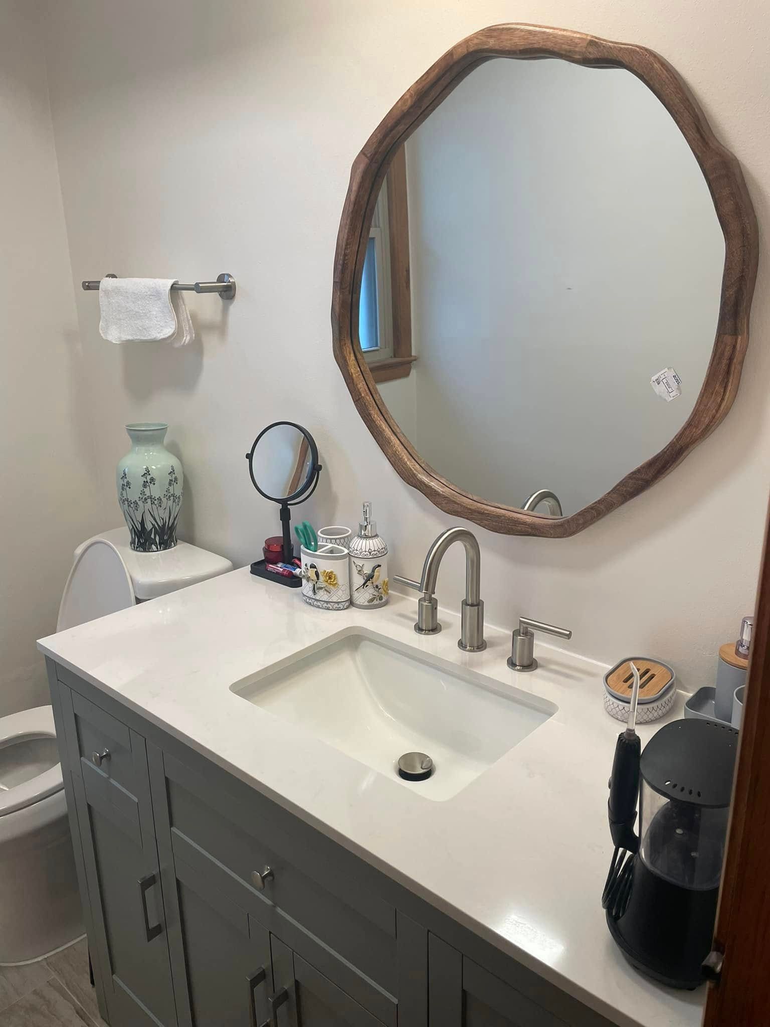 Bathroom vanity with white sink, round wood-framed mirror, gray cabinet, and countertop items.