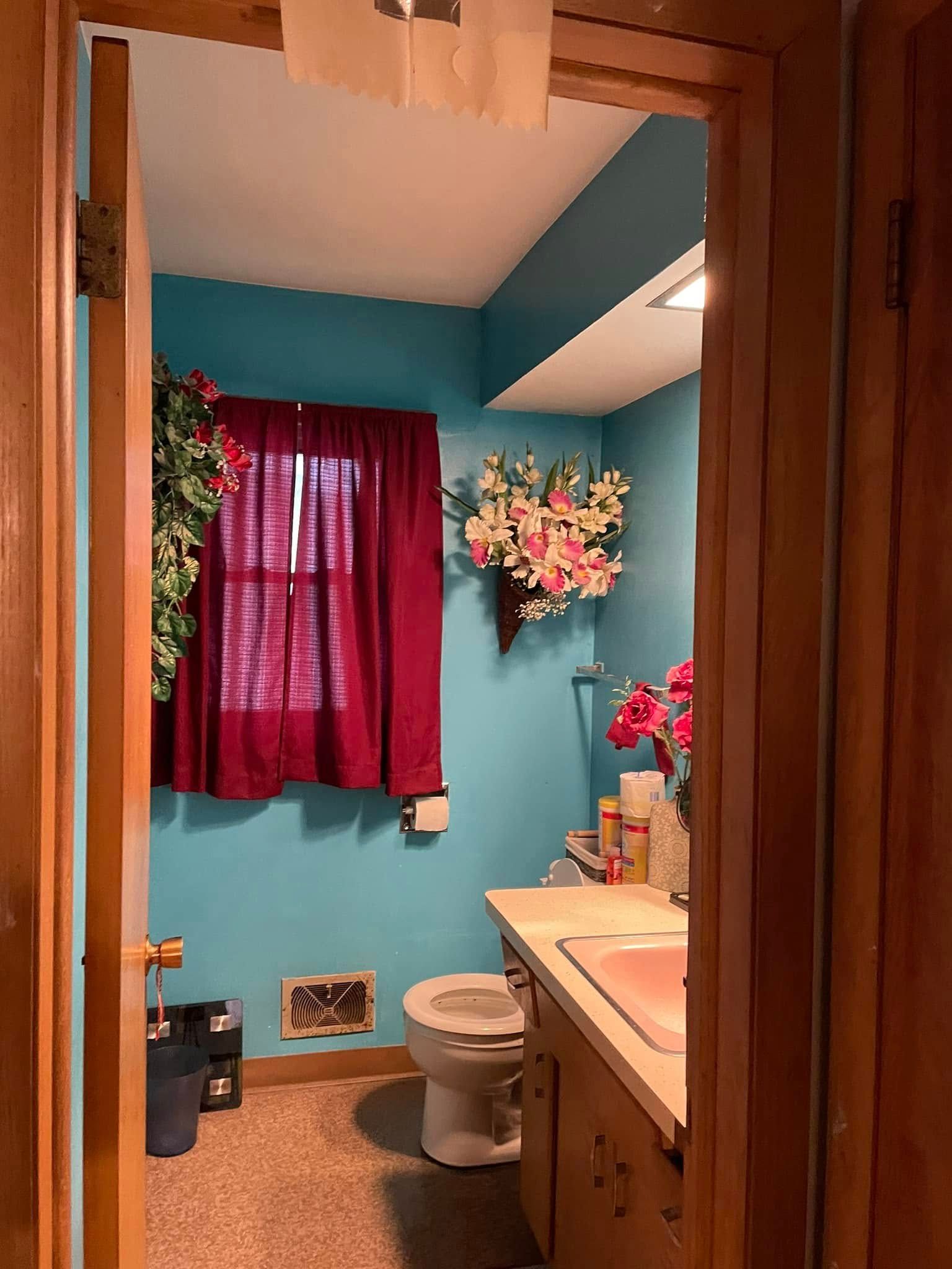 Small bathroom with teal walls, red curtain, white toilet, and sink with flowers.