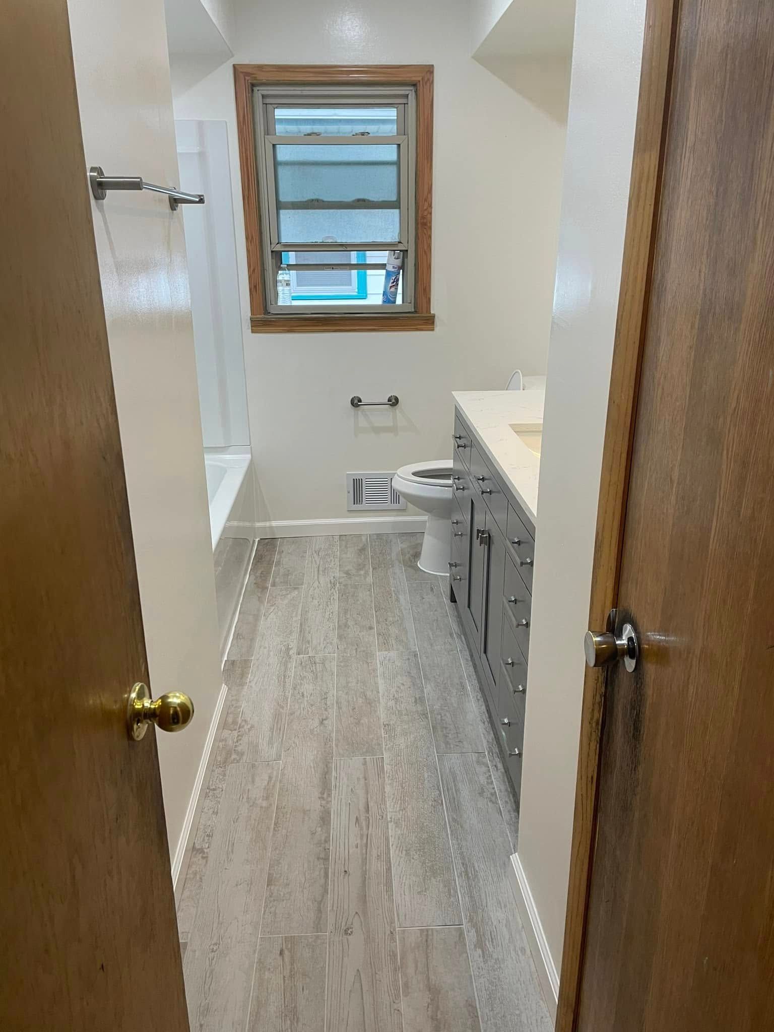 Narrow bathroom with toilet, vanity, window, and wood trim, viewed through an open door.