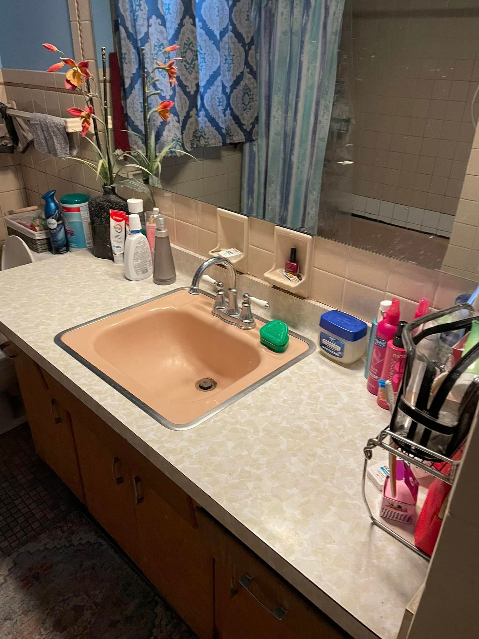 Bathroom sink vanity with tan countertop, mirror, toiletries, and blue curtains