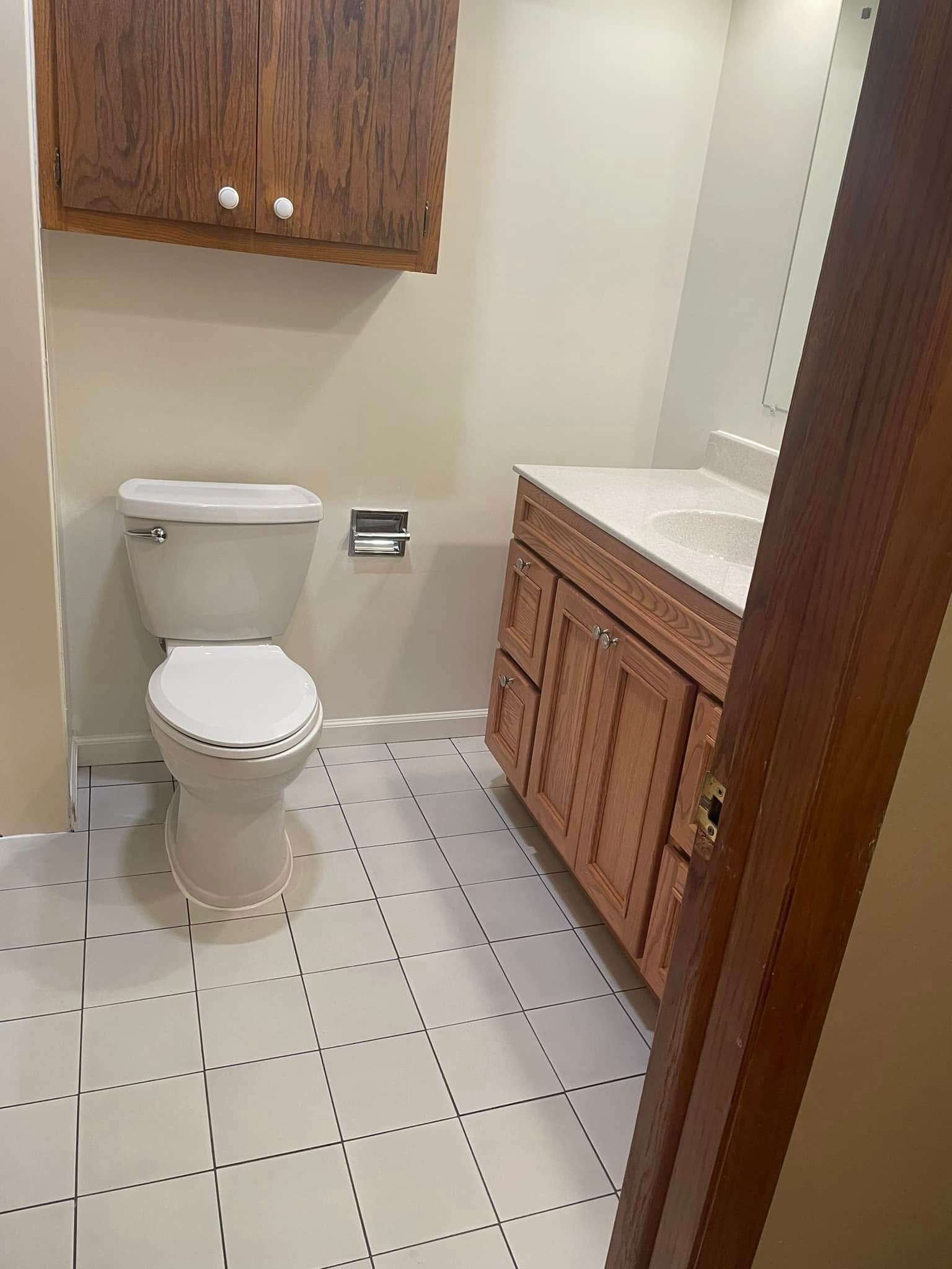 Small bathroom with a toilet, vanity sink, wood cabinets, and white tile floor.