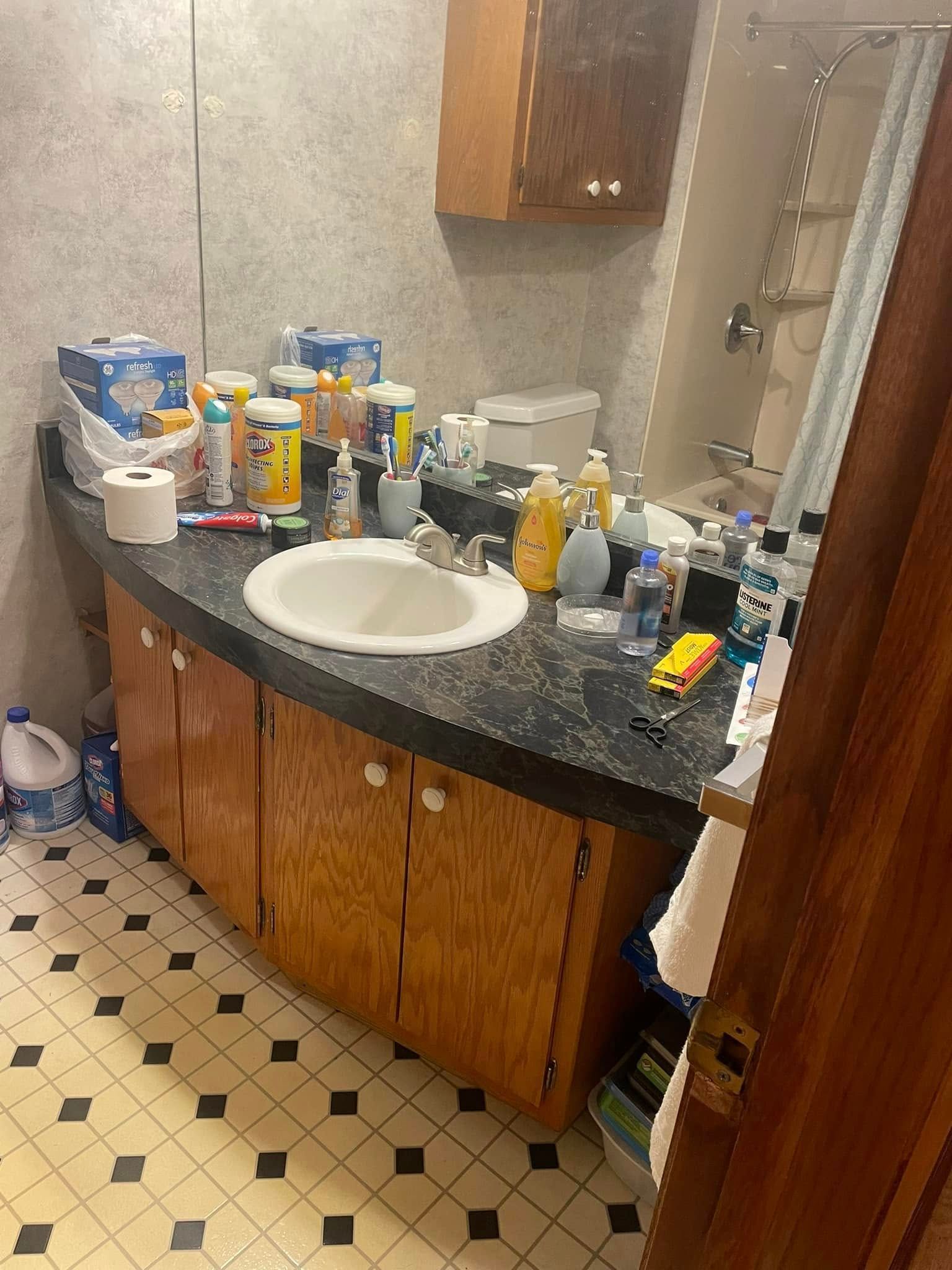 Cluttered bathroom vanity with sink, toiletries, and bottles on a dark countertop, with a mirror above.