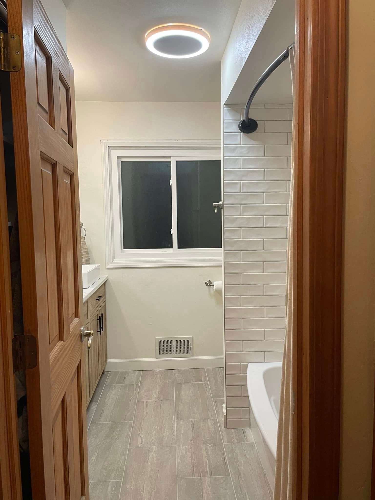 Small bathroom with wood door, white tub/shower, window, and round ceiling light.