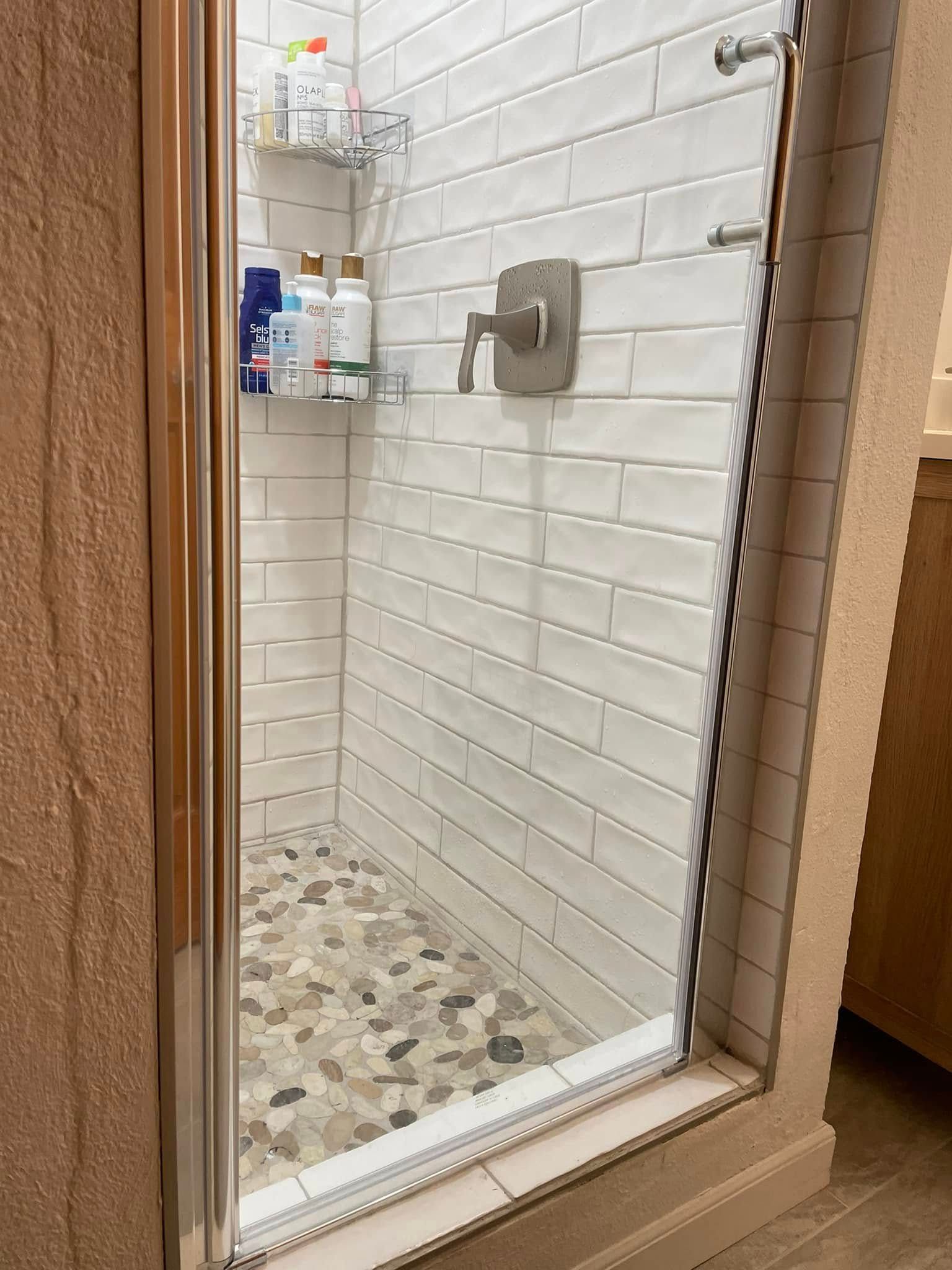 Small bathroom shower with white subway tile walls and mosaic pebble floor behind a glass door.