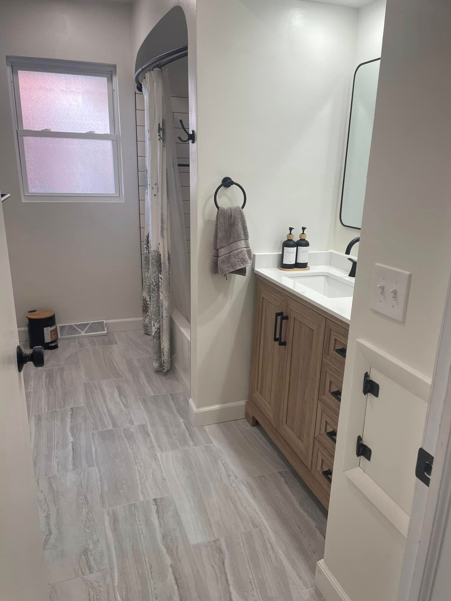 Small bathroom with gray tile floor, white walls, vanity, mirror, towel ring, and window with frosted glass.