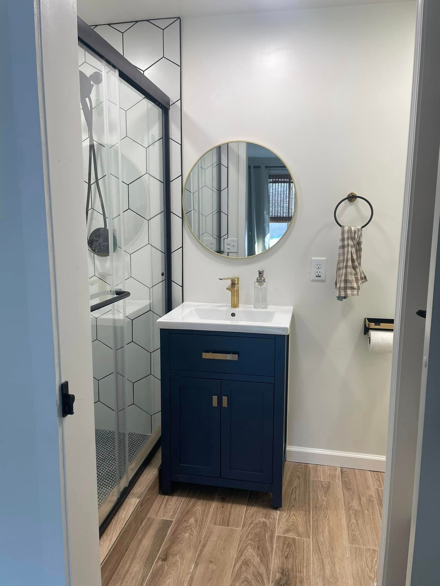 Small bathroom with a navy vanity, round mirror, glass shower, and wood-look floor