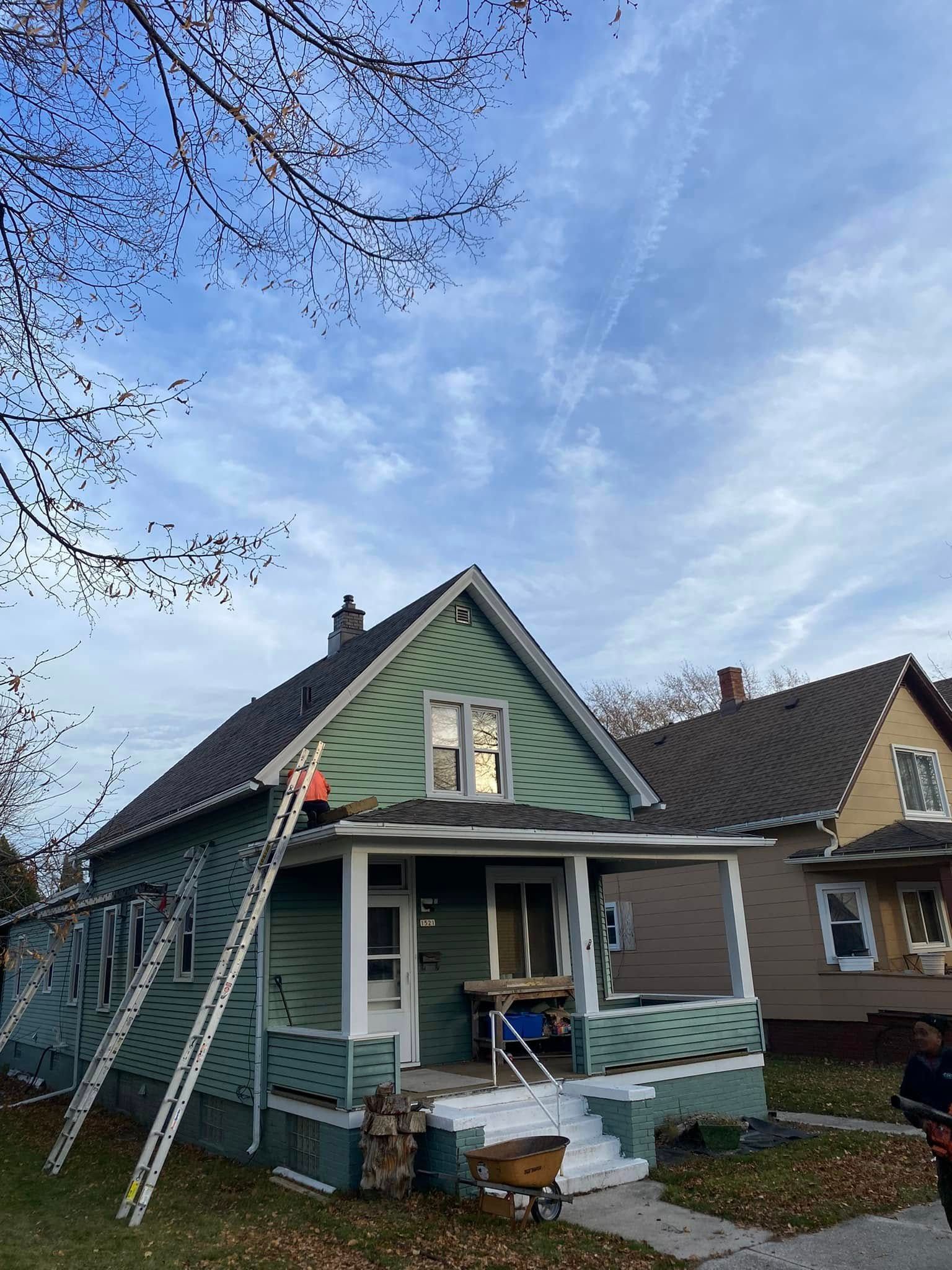 Green house with front porch and ladder, set under a blue sky with tree branches overhead