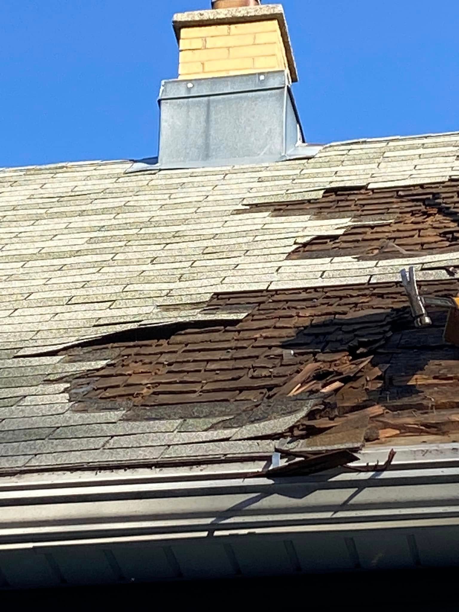 Damaged shingle roof with a large hole near the ridge and a chimney above it