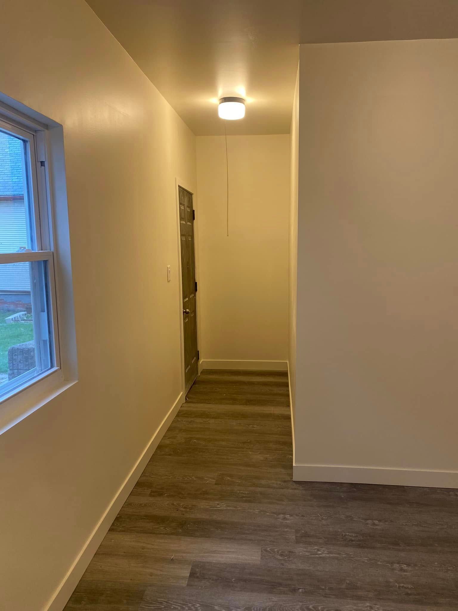 Narrow hallway with beige walls, wood-look floor, ceiling light, and a window on the left.